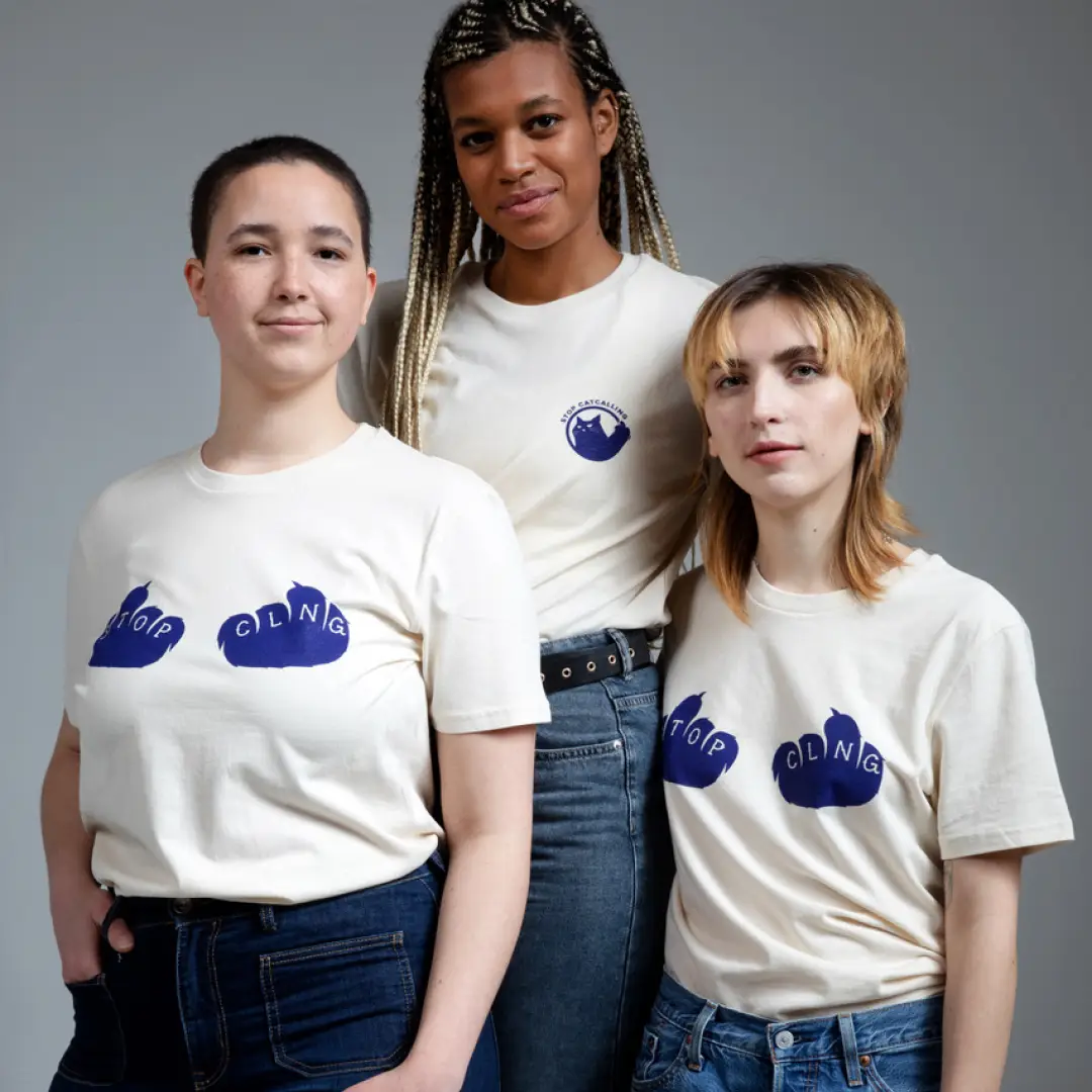 Drei Personen in wei&szlig;en T-Shirts mit dunkelblauen Aufdrucken: Zwei tragen Schriftz&uuml;ge &uuml;ber Katzenpfoten mit &sbquo;STOP&lsquo; und &sbquo;CLNG&lsquo;, eine tr&auml;gt das &sbquo;Stop Catcalling&lsquo;-Logo.