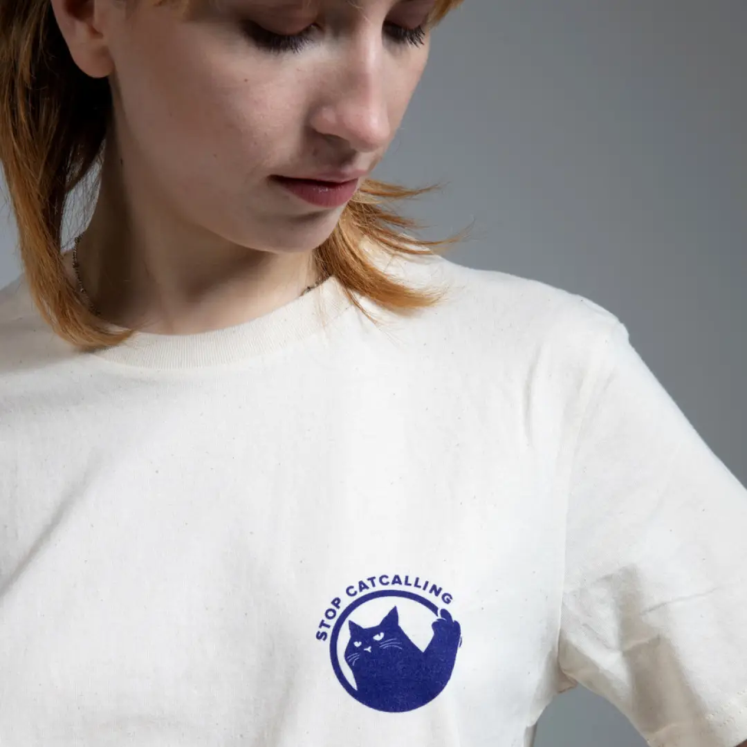 Person in wei&szlig;em T-Shirt mit blauem Logo, das eine Katze zeigt und den Schriftzug &sbquo;Stop Catcalling'