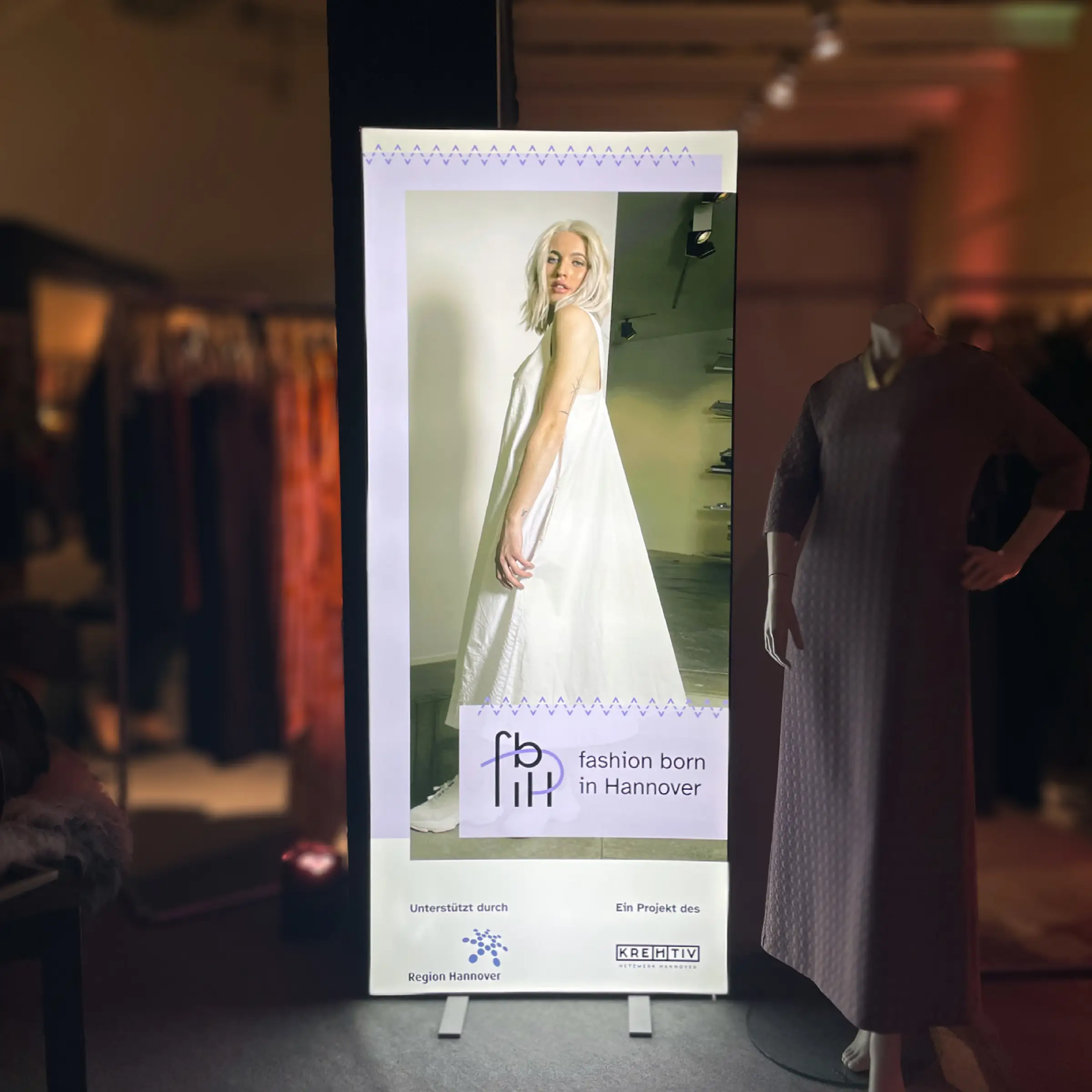 Foto eines hochformatigen Roll-Up-Banners, welches erleuchtet ist. Es zeigt ein Layout mit einer blonden Frau, welche ein wei&szlig;es Kleid tr&auml;gt und seitlich zur Kamera posiert. Dar&uuml;ber liegt eine helle Fl&auml;che mit dem fbiH Logo.